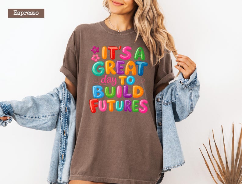 Pode incluir: Camiseta cor de caf&eacute; com a frase "IT'S A GREAT DAY TO BUILD FUTURES" em letras coloridas de bolhas. A camiseta tem mangas enroladas e &eacute; usada com uma jaqueta jeans azul.