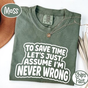 May include: A moss green short-sleeve t-shirt featuring the white text "TO SAVE TIME LET'S JUST ASSUME I'M NEVER WRONG" printed in a bold, rounded font. The shirt has a crew neckline and a Comfort Colors brand label inside the collar.