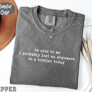 Może przedstawiać: Szara koszulka Comfort Colors z białym napisem "be nice to me I probably lost an argument to a toddler today". Złoty naszyjnik z wisiorkiem jest ułożony na koszulce.