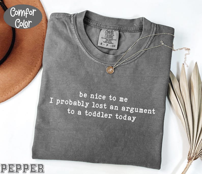 Może przedstawiać: Szara koszulka Comfort Colors z białym napisem "be nice to me I probably lost an argument to a toddler today". Złoty naszyjnik z wisiorkiem jest ułożony na koszulce.