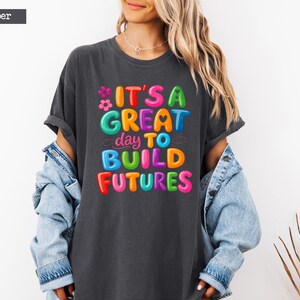 Pode incluir: Uma camiseta cinza escuro com o texto "IT'S A GREAT day TO BUILD FUTURES" em letras coloridas. A camiseta &eacute; usada com uma jaqueta jeans azul clara.