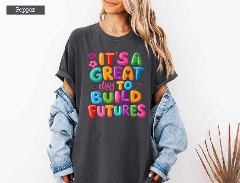 Pode incluir: Uma camiseta cinza escuro com o texto "IT'S A GREAT day TO BUILD FUTURES" em letras coloridas. A camiseta &eacute; usada com uma jaqueta jeans azul clara.