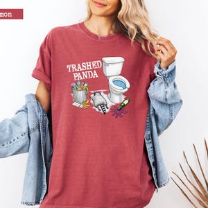 K&ouml;nnte beinhalten: Karmesinrotes T-Shirt mit dem Text "Trashed Panda" und einer Grafik einer Toilette, eines M&uuml;lleimers und eines Waschb&auml;ren. Das Shirt hat eine lockere Passform und hochgekrempelte &Auml;rmel.