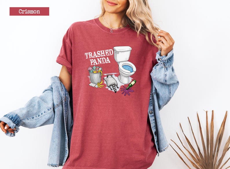 K&ouml;nnte beinhalten: Karmesinrotes T-Shirt mit dem Text "Trashed Panda" und einer Grafik einer Toilette, eines M&uuml;lleimers und eines Waschb&auml;ren. Das Shirt hat eine lockere Passform und hochgekrempelte &Auml;rmel.