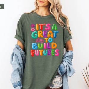 Pode incluir: Uma camiseta verde musgo com o texto "IT'S A GREAT day TO BUILD FUTURES" em letras de bolhas coloridas. A camiseta tem um corte folgado e mangas curtas. O texto inclui flores e cora&ccedil;&otilde;es.