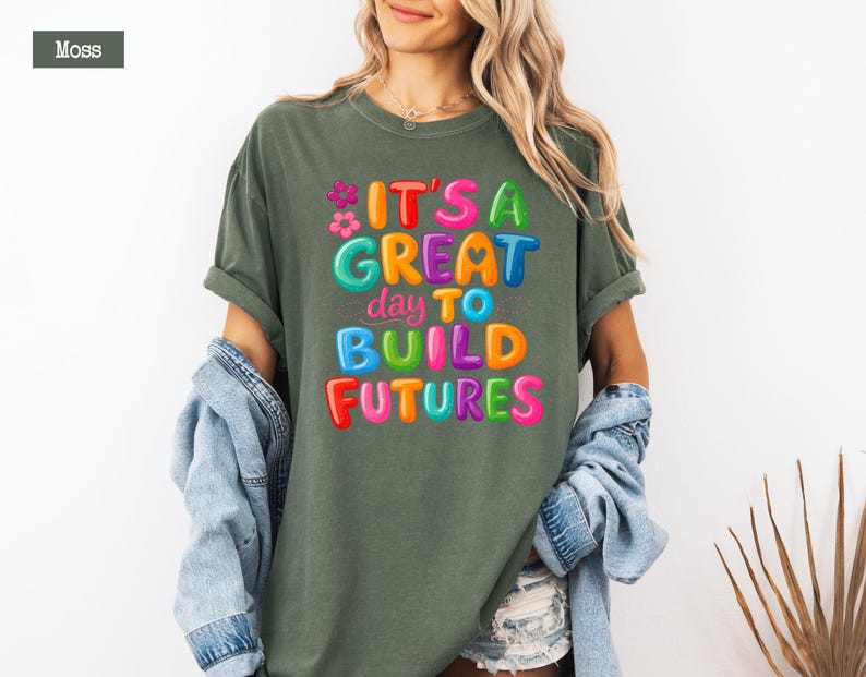 Pode incluir: Uma camiseta verde musgo com o texto "IT'S A GREAT day TO BUILD FUTURES" em letras de bolhas coloridas. A camiseta tem um corte folgado e mangas curtas. O texto inclui flores e cora&ccedil;&otilde;es.