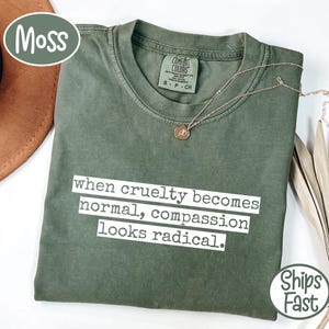 Peut inclure: Un t-shirt vert mousse avec le texte "when cruelty becomes normal, compassion looks radical" en blanc. Un collier doré avec un pendentif repose sur le t-shirt. Le t-shirt est plié et présenté avec un chapeau et une plante séchée.