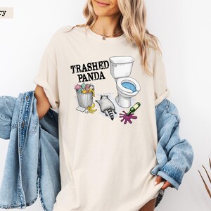 K&ouml;nnte beinhalten: Elfenbeinfarbenes T-Shirt mit dem Text "Trashed Panda" &uuml;ber einer Grafik eines Waschb&auml;ren, einer Toilette und einem M&uuml;lleimer. Der Waschb&auml;r befindet sich neben einer Toilette mit blauem Wasser, einem M&uuml;lleimer mit bunten Gegenst&auml;nden und einer versch&uuml;tteten Weinflasche.