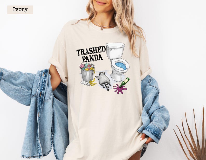 K&ouml;nnte beinhalten: Elfenbeinfarbenes T-Shirt mit dem Text "Trashed Panda" &uuml;ber einer Grafik eines Waschb&auml;ren, einer Toilette und einem M&uuml;lleimer. Der Waschb&auml;r befindet sich neben einer Toilette mit blauem Wasser, einem M&uuml;lleimer mit bunten Gegenst&auml;nden und einer versch&uuml;tteten Weinflasche.
