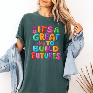 Pode incluir: T-shirt verde escuro com o texto "IT'S A GREAT day TO BUILD FUTURES" em letras de bolhas coloridas. A camisa tem um corte folgado e mangas curtas. Uma jaqueta jeans azul claro est&aacute; sobre os ombros.