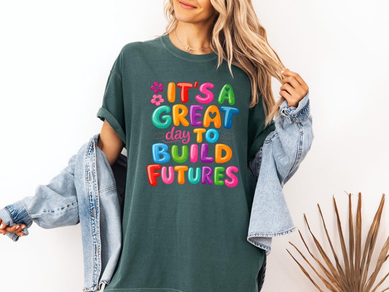 Pode incluir: T-shirt verde escuro com o texto "IT'S A GREAT day TO BUILD FUTURES" em letras de bolhas coloridas. A camisa tem um corte folgado e mangas curtas. Uma jaqueta jeans azul claro est&aacute; sobre os ombros.