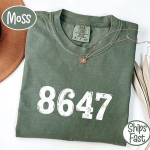 Puede incluir: Una camiseta verde musgo con el número "8647" impreso en blanco. La camiseta tiene cuello redondo y mangas cortas. Un collar dorado con un pequeño colgante está drapeado sobre la camiseta. La camiseta está doblada sobre una superficie blanca.