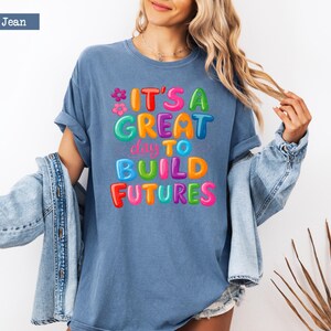 Pode incluir: Camiseta azul jeans com a frase "IT'S A GREAT day TO BUILD FUTURES" em letras coloridas. A camiseta &eacute; combinada com uma jaqueta jeans azul clara, criando um visual casual.