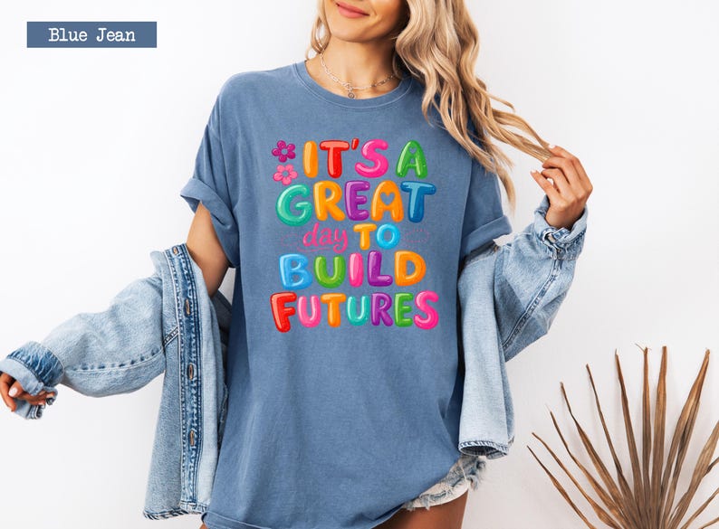 Pode incluir: Camiseta azul jeans com a frase "IT'S A GREAT day TO BUILD FUTURES" em letras coloridas. A camiseta &eacute; combinada com uma jaqueta jeans azul clara, criando um visual casual.