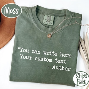 Peut inclure: Un t-shirt vert mousse avec le texte "You can write here Your custom text" en blanc. Un collier en or avec un pendentif est sur le t-shirt. Le t-shirt est plié et prêt à être porté.