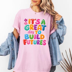 Pode incluir: Camiseta rosa claro com o texto "IT'S A GREAT day TO BUILD FUTURES" em letras coloridas. O design inclui flores e nuvens. A camiseta &eacute; usada com uma jaqueta jeans.