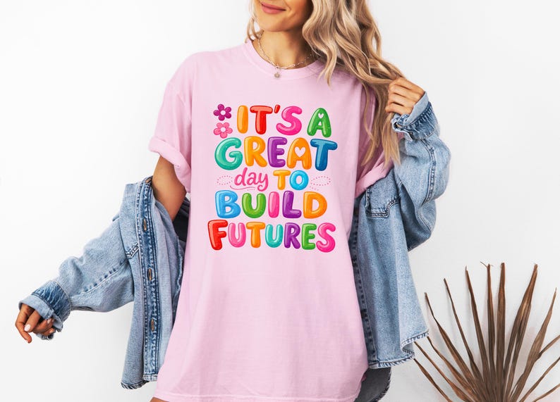 Pode incluir: Camiseta rosa claro com o texto "IT'S A GREAT day TO BUILD FUTURES" em letras coloridas. O design inclui flores e nuvens. A camiseta &eacute; usada com uma jaqueta jeans.