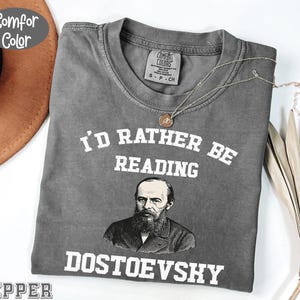 Puede incluir: Una camiseta gris Comfort Colors con el texto blanco "I'd Rather Be Reading DOSTOEVSKY". La camiseta presenta un retrato de Fiódor Dostoievski. Un collar dorado está drapeado sobre la camiseta.