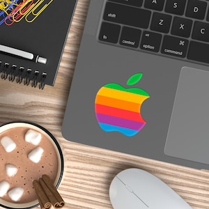 May include: A gray laptop featuring a rainbow-colored apple logo sticker on its surface, positioned next to a mug of hot cocoa with marshmallows and cinnamon sticks, a black spiral notebook with colorful paper clips, and a white computer mouse.