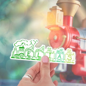 May include: A green and white train with the name "ELIAS" spelled out on the cars. The train is made of paper and is being held by a hand. The train is in front of a red toy train.