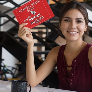 May include: A woman holds a red graduation card with the text "Illinois State Grad Congrats! Abigail Class of 2023 Agriculture".