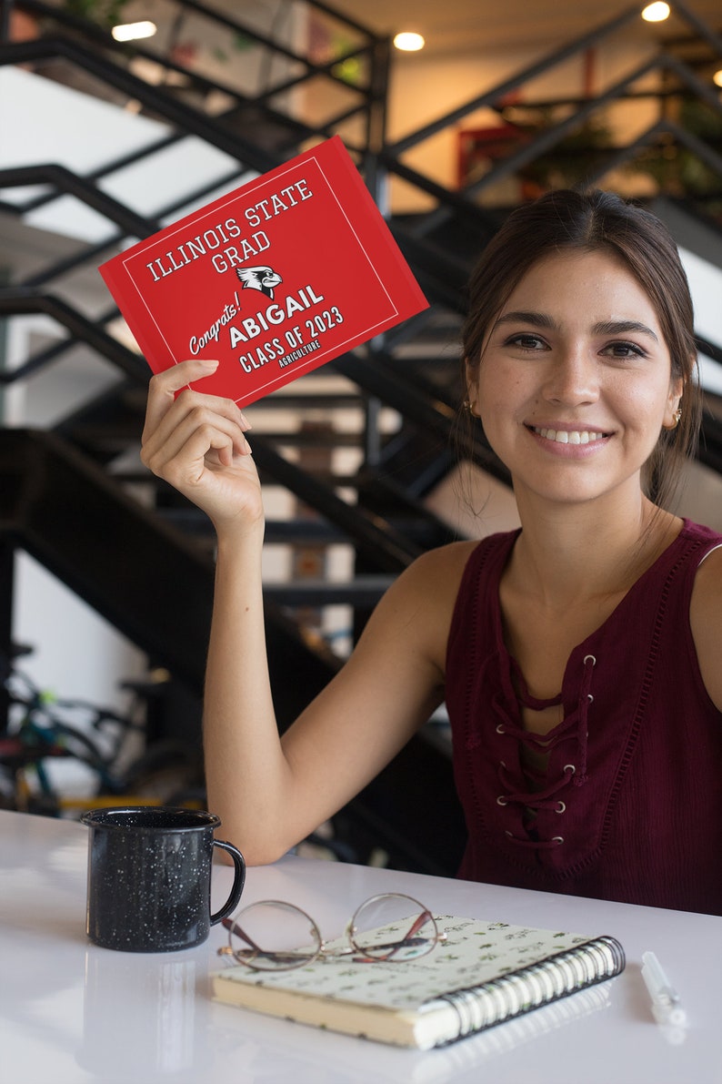 May include: A woman holds a red graduation card with the text "Illinois State Grad Congrats! Abigail Class of 2023 Agriculture".