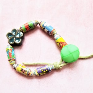 以下が含まれることがあります： 漫画のデザインが施された、カラフルな円筒形のビーズを使用した手作りのブレスレット。緑色の球形のビーズと、ターコイズブルーの花の形をしたチャームが含まれています。ブレスレットは、薄い緑色のコードに通されています。