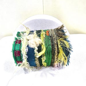 May include: A handcrafted textile art piece with a cylindrical shape. The artwork features a patchwork design with various fabrics in shades of green, blue, and gold, accented with textured yarn and fringe. The piece is set against a white background.