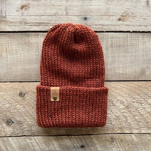May include: A rust-colored knitted beanie with a brown leather label that says "yarn & folk".