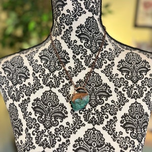 May include: A copper chain necklace with a large, raw, green and brown stone pendant. The stone is irregularly shaped and has a natural, earthy appearance. The necklace is displayed on a black and white patterned mannequin.