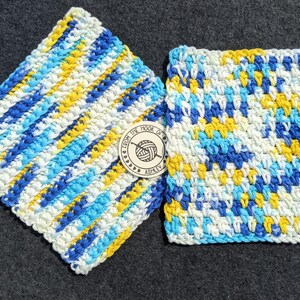 May include: Two handmade, square crochet washcloths in shades of blue, yellow, and white. One cloth has diagonal stripes, and the other has a textured pattern. A small wooden tag with the text "From the Hook of Ashley" is between them.