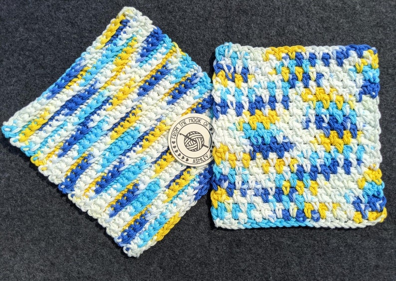 May include: Two handmade, square crochet washcloths in shades of blue, yellow, and white. One cloth has diagonal stripes, and the other has a textured pattern. A small wooden tag with the text "From the Hook of Ashley" is between them.