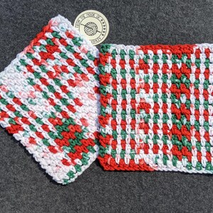 May include: Two handmade, square crochet washcloths. The washcloths feature a repeating pattern of red, green, and white yarn. One washcloth is angled, and the other is flat. A circular tag with text is visible.