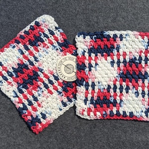 May include: Two square, handmade crochet washcloths. The washcloths are a mix of red, white, and navy blue yarn. A small, round tag with the text "From the Hook of Ashley" is attached to one of the washcloths.