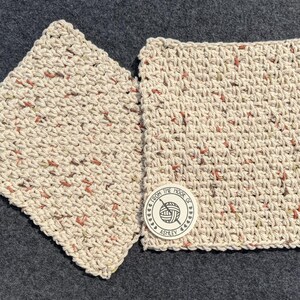 May include: Two square, hand-crocheted washcloths in a neutral beige color with flecks of red and green. One washcloth is slightly angled, and the other has a circular tag that reads "From the Hook of Ashley" with a yarn and hook logo.