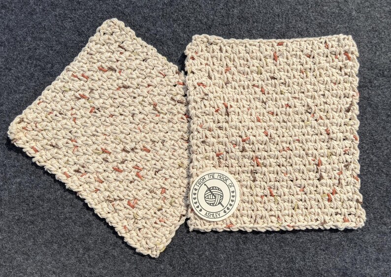 May include: Two square, hand-crocheted washcloths in a neutral beige color with flecks of red and green. One washcloth is slightly angled, and the other has a circular tag that reads "From the Hook of Ashley" with a yarn and hook logo.