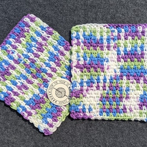May include: Two handmade crochet washcloths in a colorful pattern of blue, purple, green, and white. The washcloths are square and have a textured stitch. A wooden tag with the text "From the Hook of Ashley" is also visible.