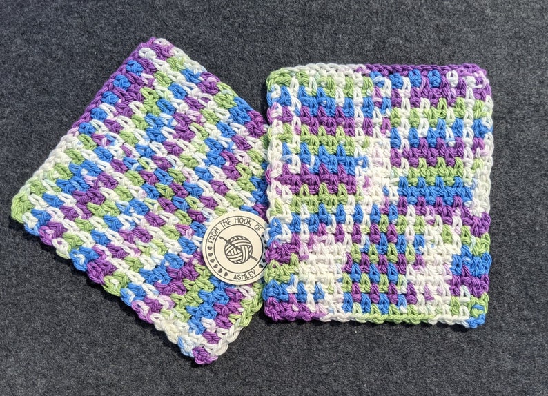 May include: Two handmade crochet washcloths in a colorful pattern of blue, purple, green, and white. The washcloths are square and have a textured stitch. A wooden tag with the text "From the Hook of Ashley" is also visible.