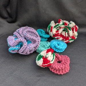 May include: A collection of handmade crochet scrunchies in various colors. The scrunchies include shades of purple, blue, green, red, white, and pink. The scrunchies are made of soft yarn and are suitable for all hair types.