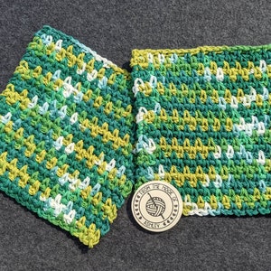May include: Two square, hand-crocheted washcloths in shades of green, yellow, and white. The washcloths are made with a textured stitch and are displayed on a gray surface. A wooden tag with the text "From the Hook of Ashley" is also present.