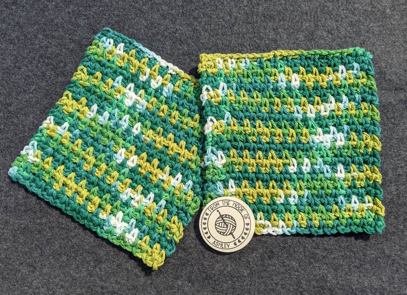 May include: Two square, hand-crocheted washcloths in shades of green, yellow, and white. The washcloths are made with a textured stitch and are displayed on a gray surface. A wooden tag with the text "From the Hook of Ashley" is also present.