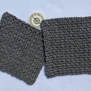 May include: Two square, gray crocheted washcloths are displayed on a white surface. The washcloths are made of a textured yarn and have a dense, tight stitch pattern. A small, circular tag with text and a yarn ball logo is visible.