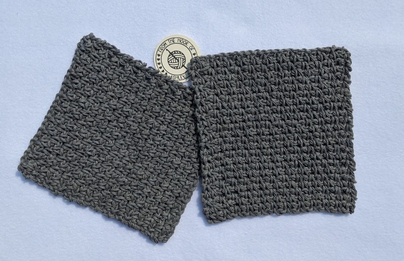 May include: Two square, gray crocheted washcloths are displayed on a white surface. The washcloths are made of a textured yarn and have a dense, tight stitch pattern. A small, circular tag with text and a yarn ball logo is visible.