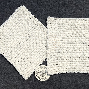 May include: Two off-white, square crocheted washcloths are displayed on a dark gray surface. The washcloths have a textured, woven appearance. A small, circular tag with text and a yarn ball graphic is visible.