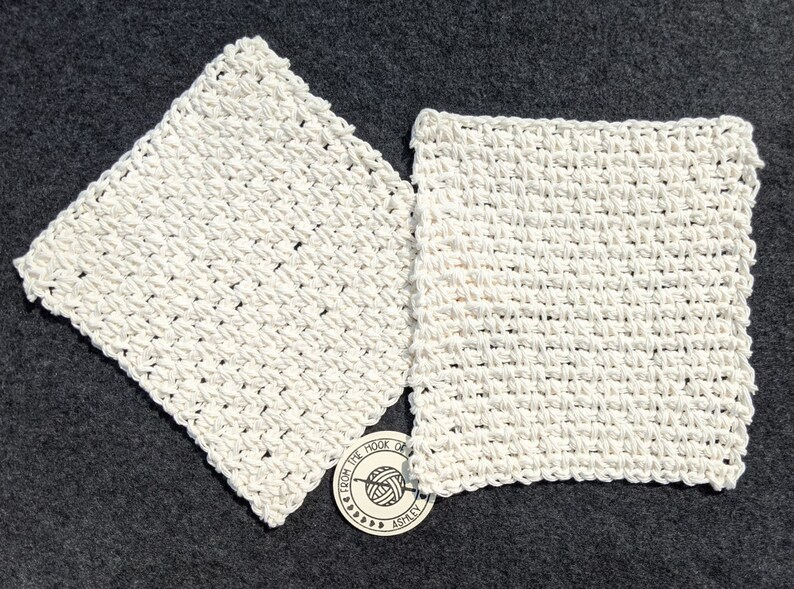 May include: Two off-white, square crocheted washcloths are displayed on a dark gray surface. The washcloths have a textured, woven appearance. A small, circular tag with text and a yarn ball graphic is visible.