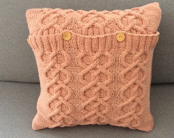 May include: A soft, peach-colored knitted pillow with a cable knit pattern and two wooden buttons.