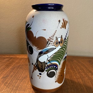 May include: A white ceramic vase with a blue rim and a hand-painted design of a bird with brown, green, and blue accents.
