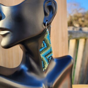 May include: A beaded earring with a diamond shape. The earring features a pattern of blue, green, and black beads. The earring is hanging from a silver hook. The earring is displayed on a black mannequin.