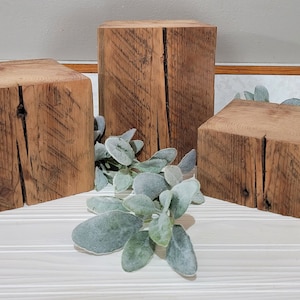 May include: Three rustic wooden blocks of varying heights, arranged on a white surface with faux greenery. The blocks are square or rectangular, with visible wood grain and natural cracks, showcasing a weathered, aged appearance. The blocks are likely used for display or decor.