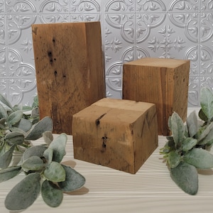 May include: Three rectangular wooden blocks of varying heights with a natural brown finish, visible wood grain, and small knots. The blocks are arranged in a cluster, accompanied by sprigs of green foliage with rounded, soft-textured leaves.